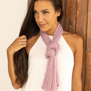 May include: A woman with long brown hair wearing a white sleeveless top and a light pink scarf tied around her neck.