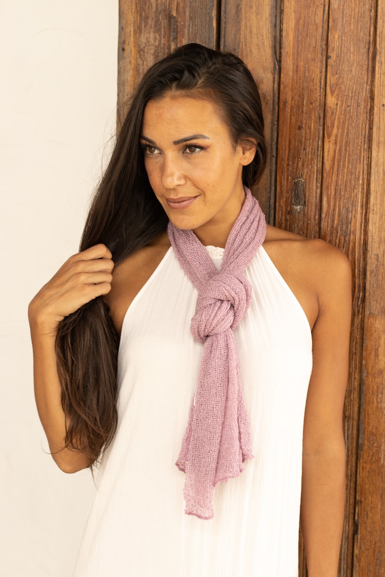 May include: A woman with long brown hair wearing a white sleeveless top and a light pink scarf tied around her neck.