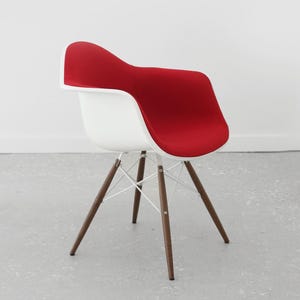 May include: A modern armchair with a white molded plastic seat and a red upholstered backrest. The chair has a wooden base with dark brown legs and white metal supports. The design is minimalist and stylish, suitable for various interior settings.