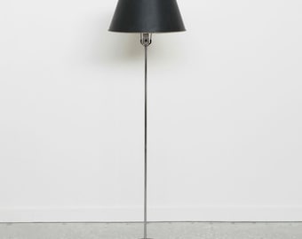 May include: A tall, slender floor lamp with a black lampshade. The lampshade is conical and sits atop a chrome pole. The base is a dark brown disc. The lamp is plugged into an electrical outlet.