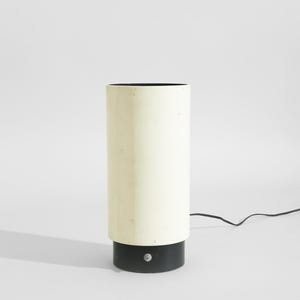 May include: A cylindrical table lamp with a cream-colored shade and a black base. The lamp has a simple, modern design and a black power cord. The lamp is approximately 12 inches tall.