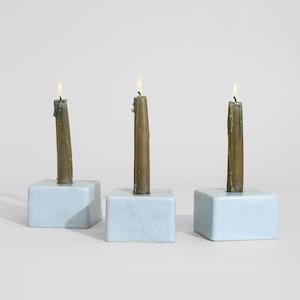 May include: Three light blue square candle holders with a speckled finish, each holding a single lit olive green taper candle. The holders are arranged in a row, featuring a smooth, matte texture and a flat top surface with a central hole.