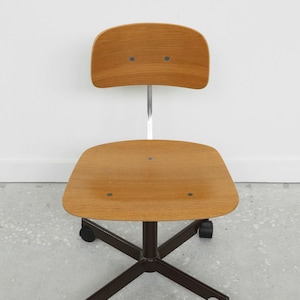 May include: A wooden office chair with a curved back and seat, mounted on a dark brown metal base with five wheels. The chair's back and seat are made of light brown wood. The chair has a chrome-colored support column.