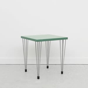 May include: A small, square, green table with six thin, silver legs. The table is on a white floor.
