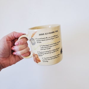 May include: Cream ceramic mug with the text "BASIC FLY FISHING TIPS" and illustrations of fishing equipment. The mug displays four fly fishing tips, including how to avoid break-offs and protect waders.