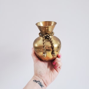 Peut inclure: Un vase en laiton doré avec un motif de corde autour du cou. Le vase a un corps rond et une large ouverture.