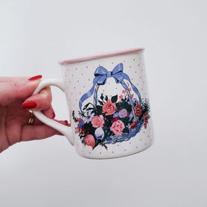 May include: White ceramic mug with a pink rim and a floral design. The design features a blue ribbon tied around a basket filled with pink, red, and white flowers. The mug has a white background with pink polka dots.