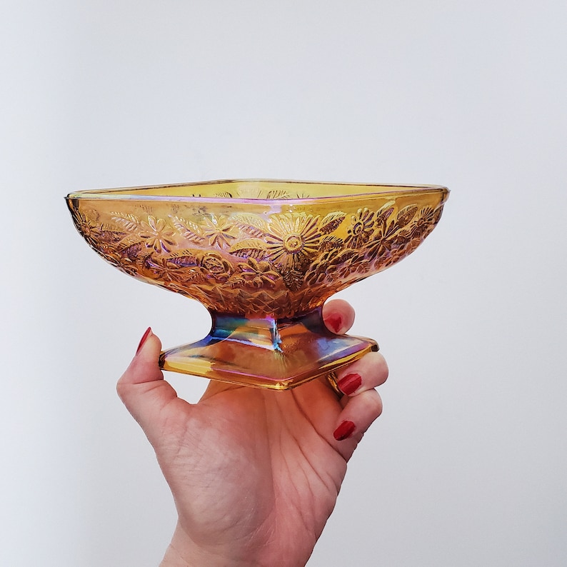May include: A vintage iridescent amber glass candy dish with a floral design. The dish has a square base and a wide, shallow bowl.