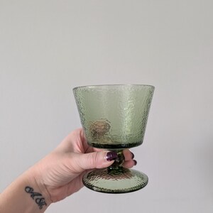 May include: A green textured glass goblet with a rounded base and a short stem. The glass has a slightly flared top and a textured surface. The goblet is held against a plain white background.