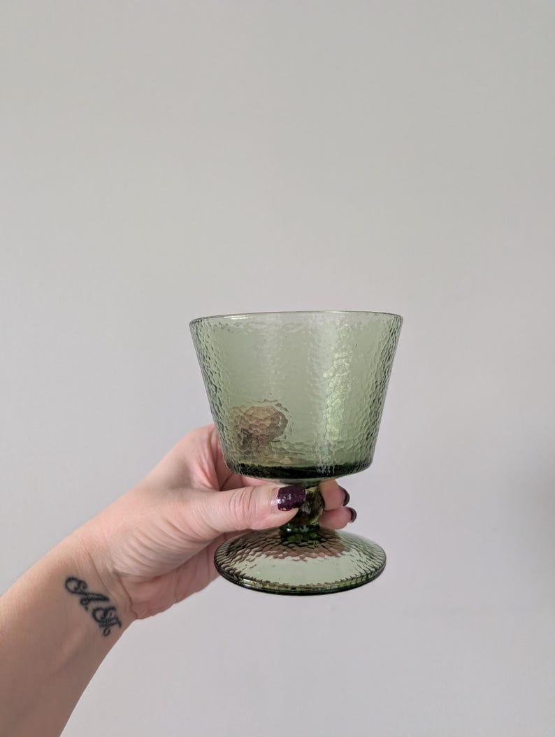 May include: A green textured glass goblet with a rounded base and a short stem. The glass has a slightly flared top and a textured surface. The goblet is held against a plain white background.