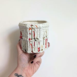 May include: A cylindrical ceramic planter with a textured, off-white exterior. The planter features vertical ridges and a speckled pattern with red and brown drips. The top rim has a rope-like design. The interior is white.