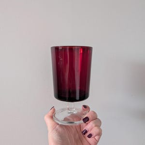 May include: A ruby red wine glass with a clear stem and base. The glass has vertical ridges and is held by a hand with dark nail polish. The glass is against a plain white background.