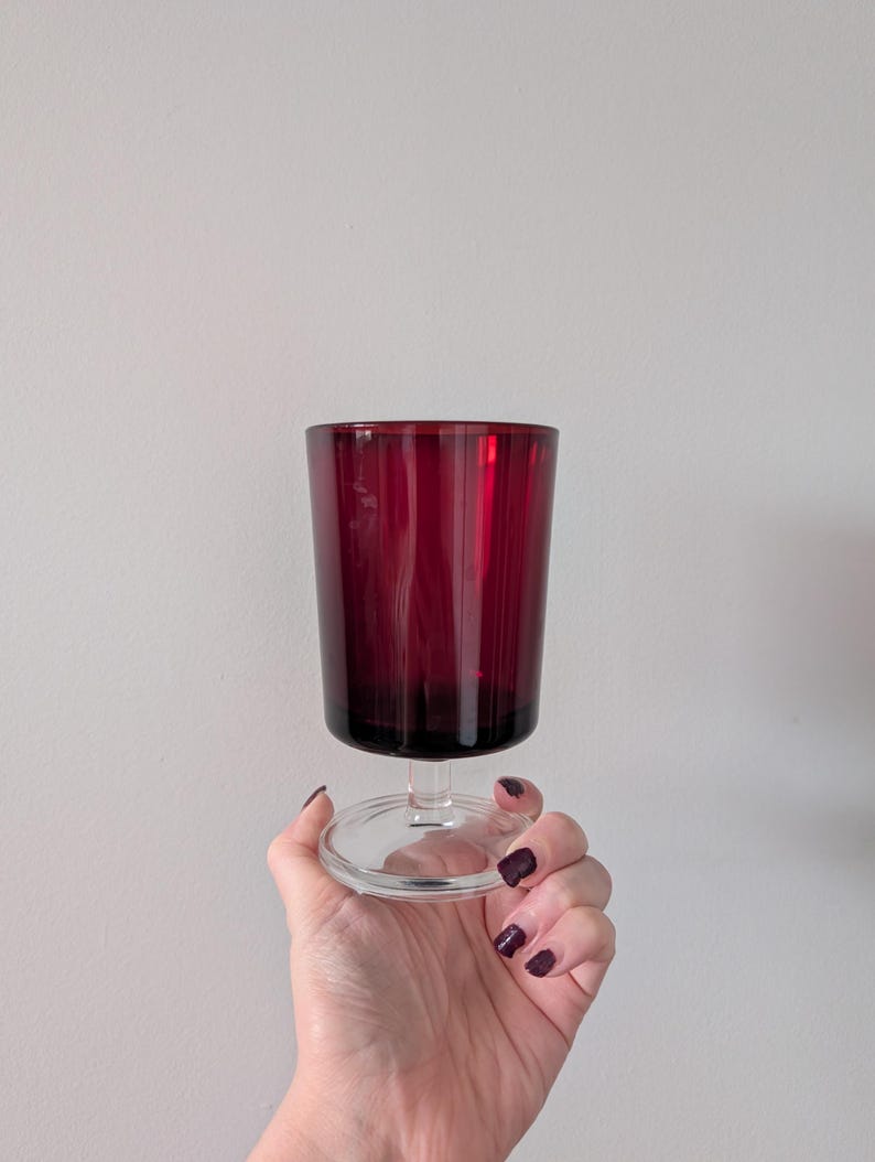 May include: A ruby red wine glass with a clear stem and base. The glass has vertical ridges and is held by a hand with dark nail polish. The glass is against a plain white background.