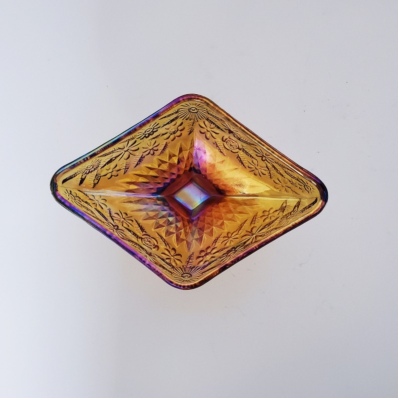 May include: A square, iridescent glass dish with a diamond pattern and floral design. The dish is a deep amber color with hints of purple and red.