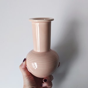 Op de afbeelding: Een lichtroze keramische vaas met een ronde basis en een cilindrische hals. De vaas heeft een geribbeld ontwerp rond de basis.