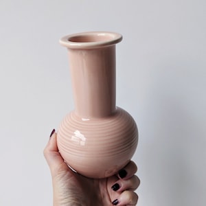 May include: A pale pink ceramic vase with a round base and a cylindrical neck. The vase has a subtle ribbed texture.
