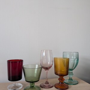 May include: A collection of five colorful drinking glasses. The glasses vary in shape and color, including red, green, pink, amber, and teal. Each glass has a unique design, with some featuring textured surfaces.
