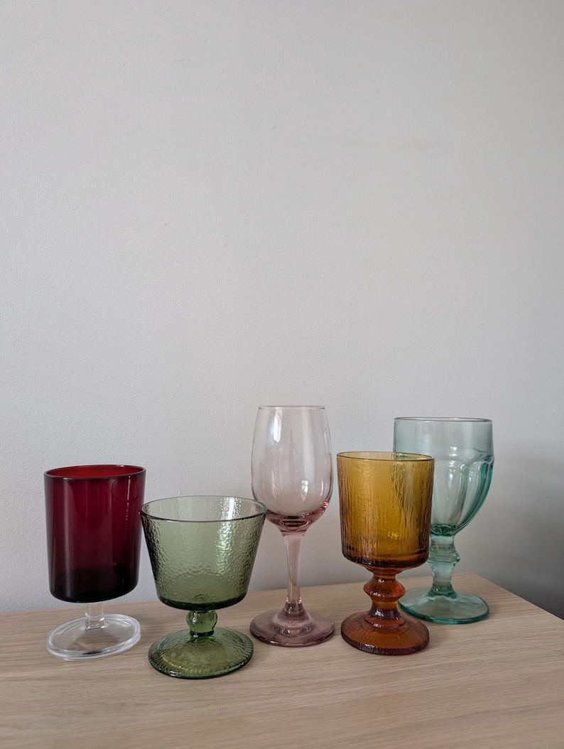May include: A collection of five colorful drinking glasses. The glasses vary in shape and color, including red, green, pink, amber, and teal. Each glass has a unique design, with some featuring textured surfaces.