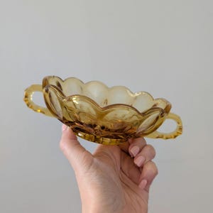 May include: Amber-colored glass bowl with a scalloped rim and two small handles. The bowl has a textured, floral-like pattern on the exterior. The bowl is held up against a plain background.