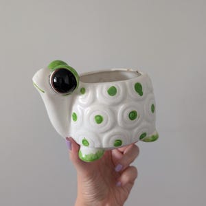May include: A white ceramic turtle-shaped planter featuring a glossy finish, a large black eye, and a shell decorated with raised circular patterns accented by green painted dots. The turtle has four small feet and an open top for planting.