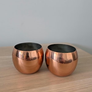 May include: Two spherical cups with a polished copper exterior finish and a dark, matte interior. The cups have a rounded, bowl-like shape with a wide opening at the top, reflecting light on their smooth, metallic surfaces.