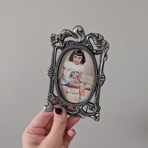 May include: An antique silver-coloured metal picture frame with an oval aperture. The frame contains a vintage photograph of a young girl. The frame features decorative scrollwork and the text "Frame for 2x3 photograph".