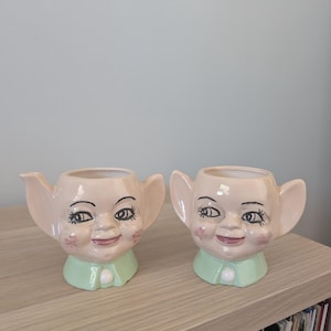 以下が含まれることがあります： 大きな耳、バラ色の頬、緑色の襟が付いた、2つのセラミック製の顔プランター。 プランターは、小さな植物や装飾的な使用に最適な、気まぐれなデザインです。 プランターは木製の棚に展示されています。