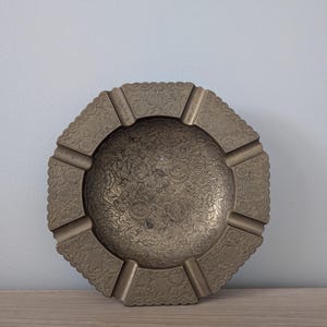 Peut inclure: Un cendrier vintage en laiton avec un motif floral complexe. Le cendrier a un centre rond avec quatre supports rectangulaires et un bord festonné décoratif. Le laiton a une patine chaude et vieillie.