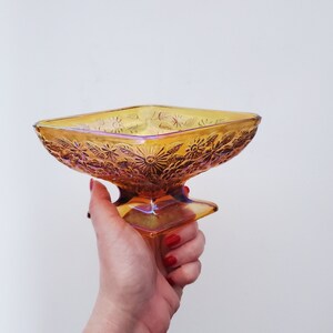 May include: A square, amber glass serving dish with a floral design. The dish has a pedestal base and is iridescent.