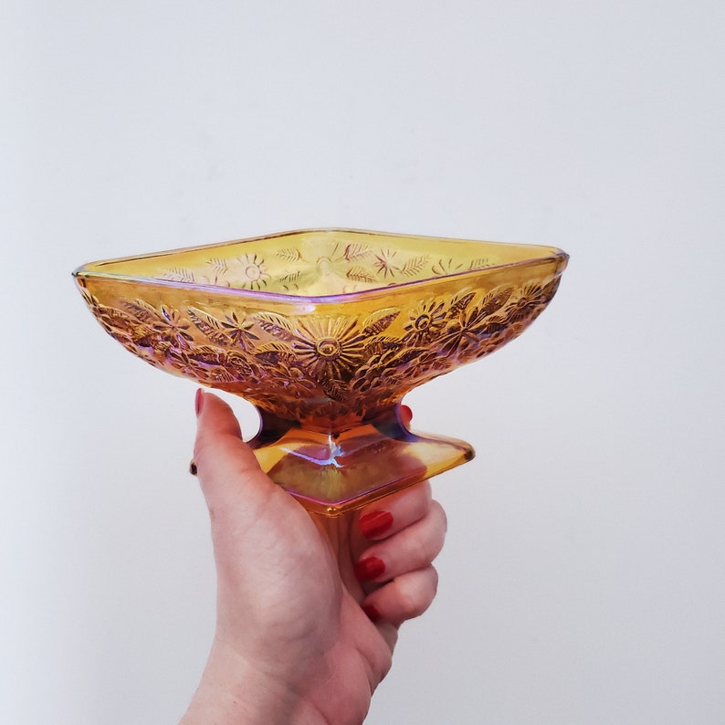 May include: A square, amber glass serving dish with a floral design. The dish has a pedestal base and is iridescent.