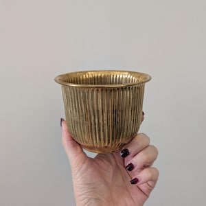 May include: A small, gold-coloured metal bowl with vertical ribbed detailing. The bowl has a rounded rim and a cylindrical shape. The bowl is held in a hand against a plain, light-coloured background. The bowl is likely used for decorative purposes.