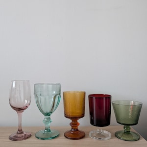 May include: Five colorful stemmed glasses in shades of pink, teal, amber, burgundy, and green are arranged on a light wood surface. The glasses vary in shape and texture, showcasing a vintage aesthetic.