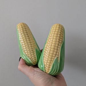 Peut inclure: Une paire de salières et poivrières en céramique en forme d'épis de maïs. Les salières et poivrières ont un motif de grains de maïs jaunes avec des feuilles vertes. Elles sont tenues dans une main sur un fond uni.