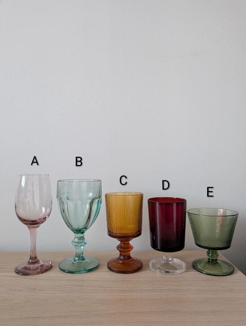 May include: Five colorful vintage-style drinking glasses. The glasses are in shades of pink, teal, amber, burgundy, and green. Each glass has a unique shape and design, perfect for a variety of beverages.