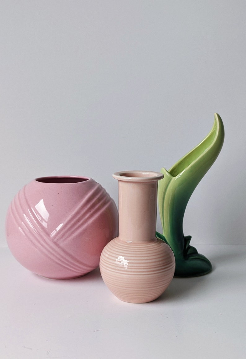 Op de afbeelding: Drie keramische vazen in roze en groen. De vaas links is een ronde, roze vaas met een geribbeld ontwerp. De vaas in het midden is een roze vaas met een rond lichaam en een smalle hals. De vaas rechts is een groene vaas met een gebogen, bladvormig ontwerp.