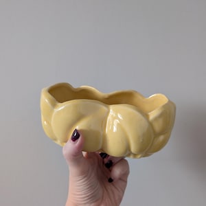 May include: A yellow ceramic bowl with a wavy rim. The bowl has a shiny surface and a curved form. The bowl is held by a hand with a tattoo on the wrist.