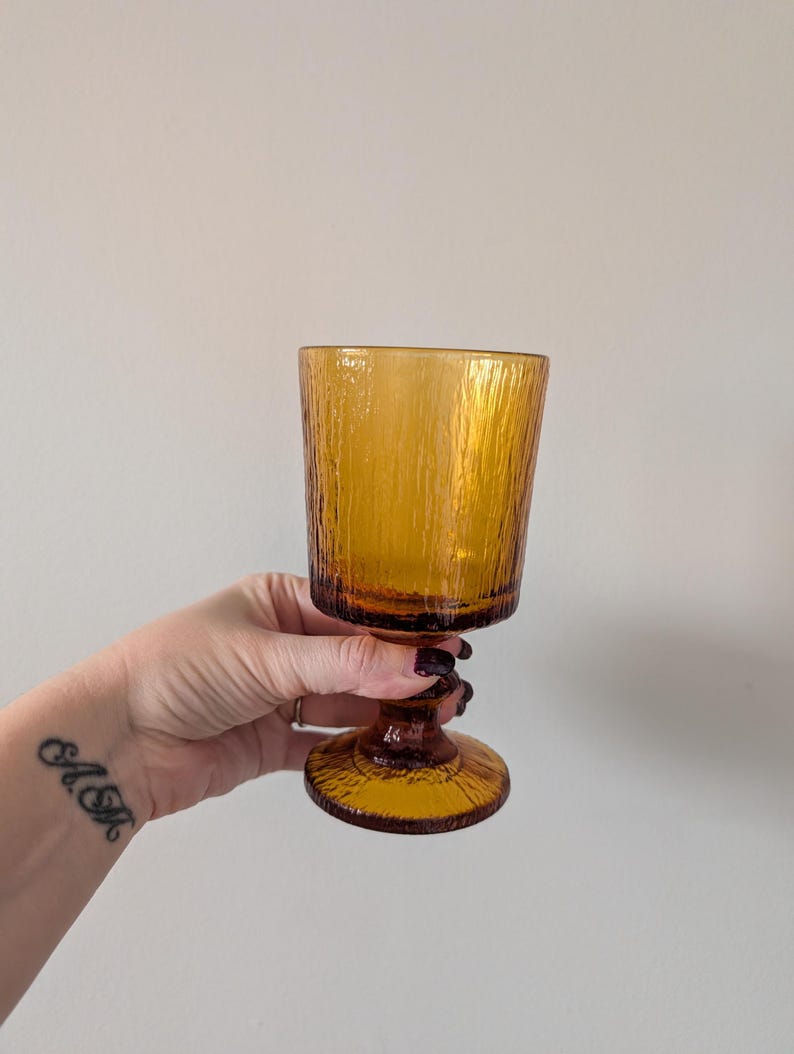 May include: Amber-colored textured glass goblet. The goblet has a textured, cylindrical bowl and a dark brown stem with a decorative, twisted design. The base is round and also textured. The glass is held in a hand.