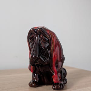 May include: A ceramic figurine of a Basset Hound dog, featuring a glossy finish with shades of red and brown. The dog is depicted in a seated position with a sad expression, showcasing long ears and a droopy face. The figurine is placed on a wooden surface.