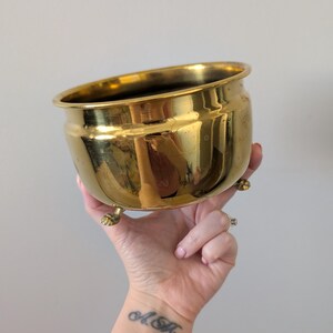 Peut inclure: Un pot en laiton doré et brillant, avec un corps arrondi et trois petits pieds. Le pot a un large bord légèrement évasé. L'intérieur semble sombre. Le pot est tenu dans une main.
