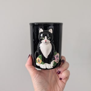 Puede incluir: Jardinera de cerámica negra con un gato blanco y negro, ojos verdes y nariz rosa. El gato está rodeado de flores de colores. La jardinera se sostiene en una mano.