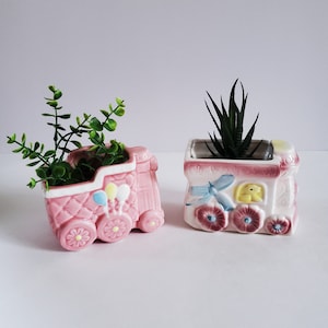 May include: Two pink ceramic planters shaped like a train engine and a train car. The train engine planter has a green plant inside and the train car planter has a green succulent inside.