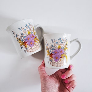 May include: Two white ceramic mugs with floral designs. Both mugs feature a basket of purple flowers with green leaves and blue forget-me-nots. One mug has a handle on the right side, the other on the left.
