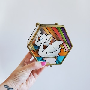 May include: A hexagonal glass trinket box with a gold frame. The box features a stained glass design of two white swans swimming in a rainbow-colored lake.
