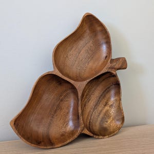 May include: A three-section wooden serving dish carved into a leaf shape with a prominent stem on the right side. The bowl features a rich, warm brown wood grain pattern with smooth, polished surfaces and deep, rounded compartments for food.