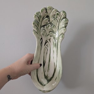 May include: A ceramic decorative platter shaped like a leafy vegetable, with a white base and green painted details. The platter is held by a hand against a grey background.