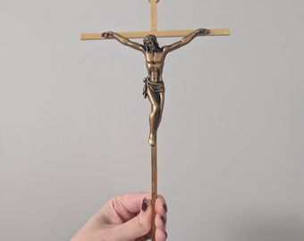 May include: A bronze-toned crucifix with a detailed figure of Jesus Christ. The cross is a simple design with a vertical post and horizontal beam. The figure is depicted with arms outstretched, and a crown of thorns. The crucifix is held by a hand.