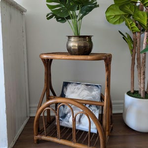May include: A vintage rattan side table with a shelf and magazine rack. A brass planter with a green plant sits on top. A framed picture is in the magazine rack. The table is made of light brown rattan.
