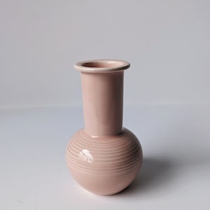Op de afbeelding: Een lichtroze keramische vaas met een ronde basis en een cilindrische hals. De vaas heeft een subtiel gestreept patroon.