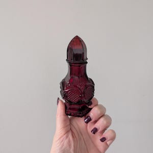 Peut inclure: Un flacon de parfum en verre rouge rubis avec un design décoratif et orné. Le flacon a un bouchon conique pointu et une base bulbeuse avec des motifs floraux en relief. Le flacon est tenu dans une main sur un fond uni.