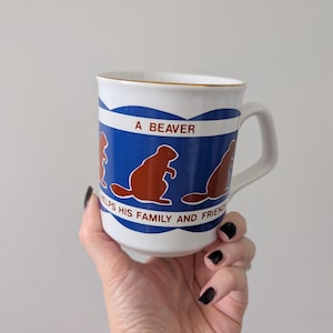 May include: White ceramic mug with a blue band featuring brown beaver illustrations and the text "A BEAVER HELPS HIS FAMILY AND FRIENDS". The mug has a gold rim and a white handle.